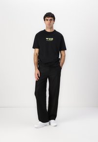 Svart kortermet T-skjorte med en limegrønn grafisk logo på brystet, kombinert med svarte bukser og hvite sneakers.
