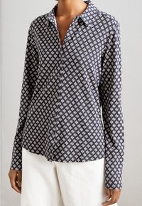 Femme portant une chemise boutonnée à manches longues à motifs sombres et un pantalon blanc, se tenant devant un arrière-plan uni.