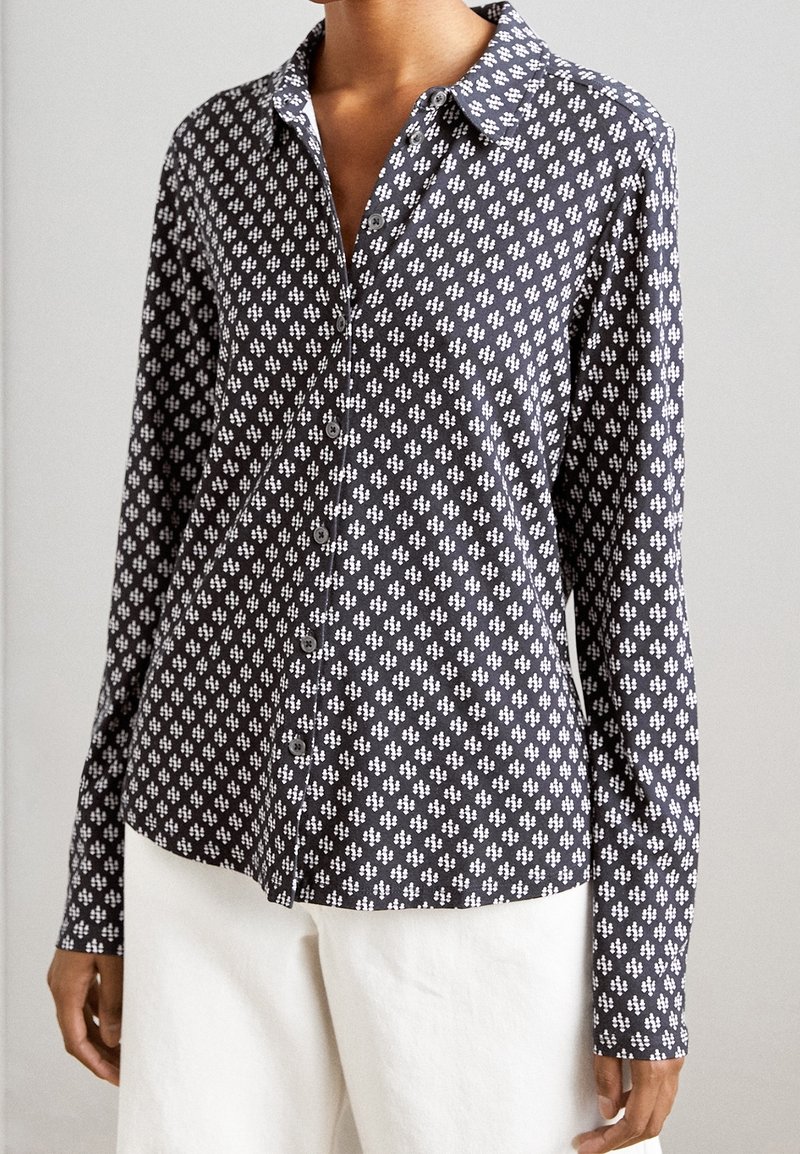 Femme portant une chemise boutonnée à manches longues à motifs sombres et un pantalon blanc, se tenant devant un arrière-plan uni.