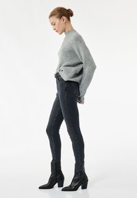 Suéter de canalé gris claro con un cuello alto, combinado con jeans skinny oscuros y botas de tobillo negras de punta afilada. Texturas suaves, diseño ajustado.