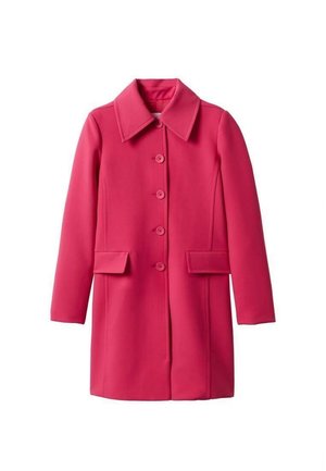 Femme portant un manteau rose vif boutonné par-dessus un crop top noir, un pantalon noir évasé avec des fentes, et des sandales à talons hauts noires avec des lanières.