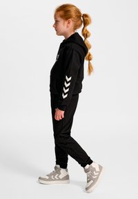 Sort hættetrøje med hvidt chevrondesign på ærmerne, kombineret med sorte joggingbukser og grå høje sneakers. Glat tekstur, afslappet pasform.