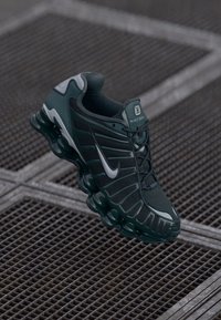 Ténis Nike Shox em preto e verde escuro com parte superior em malha texturizada, design ribbed proeminente e sola almofadada distinta.
