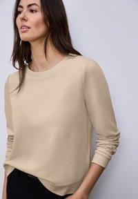 Femme portant un pull beige à manches longues avec des poignets côtelés et un pantalon noir, regardant légèrement vers la gauche sur un fond clair uni.