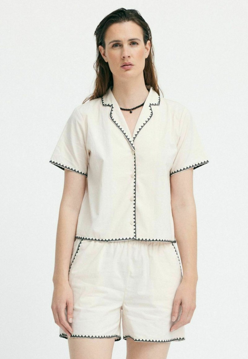 Completo a due pezzi con maniche corte in crema, caratterizzato da una camicia con bottoni e pantaloni corti, rifinito con un bordo a zigzag nero lungo i lati. Tessuto dal tocco morbido.