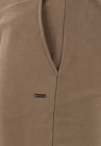 Brunt plagg med V-halsdesign, försett med en metallisk varumärkesskylt och slät, texturerad tyg. Enkel och funktionell stil.