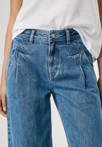 Pantaloni in denim blu chiaro con vita alta, frontale plissettato e due tasche laterali. Presentano una chiusura con bottone in metallo e cuciture a contrasto.