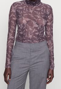 Långärmad, genomskinlig lila topp med blommönster, hög halsdesign och kroppsnära silhuett, tillsammans med grå byxor.