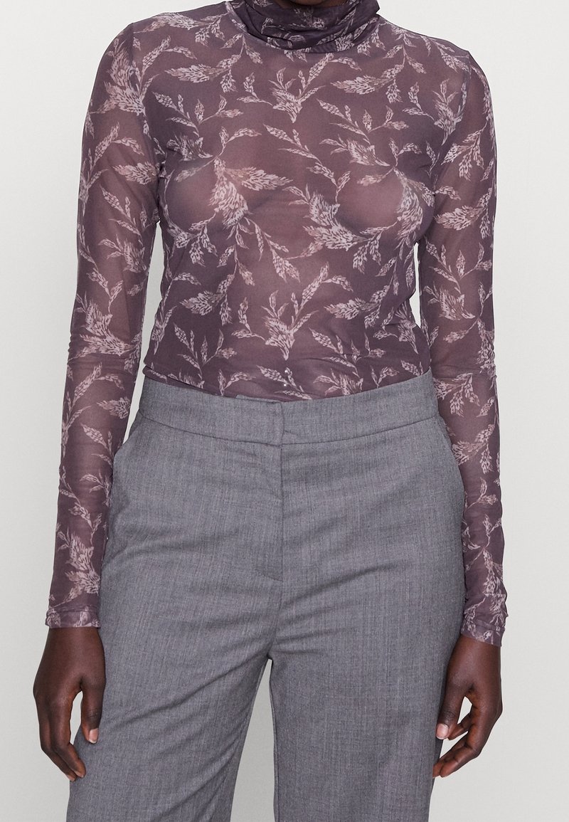 Långärmad, genomskinlig lila topp med blommönster, hög halsdesign och kroppsnära silhuett, tillsammans med grå byxor.