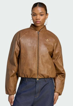 Frau trägt braune Reißverschluss-Lederjacke und blaue Jeans, mit großen Creolen und zurückgebundenem Haar, steht vor einfachem Hintergrund.