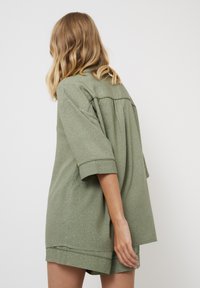 Top verde de manga corta con un corte relajado y textura estampada. Presenta un yugo en la parte posterior y mangas enrolladas, con un dobladillo bajo.