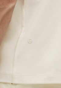 Artimiausias beige audinio vaizdas, kuriame matyti siūlės, mažas apvalus logotipas ir dalis žmogaus rankos bei liemens.