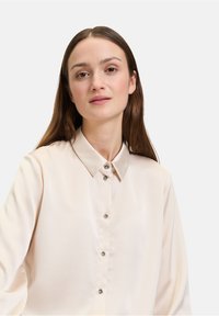 Helles beige Satin-Hemd mit Knopfleiste und Kragen, ausgestattet mit einer Vorderknopfleiste und silberfarbenen Knöpfen; lange Ärmel mit lockerer Passform.