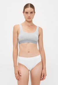 Woman standing facing forward wearing a grey sports bra and white underwear against a plain white background.