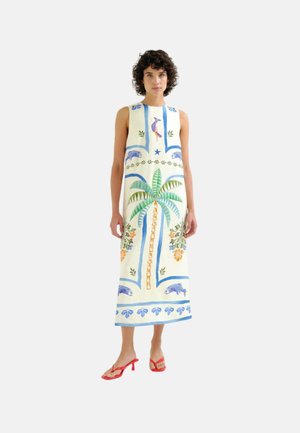 Mujer con vestido blanco sin mangas con un gran estampado de palmera y vida marina, combinado con sandalias rojas de tacón sobre un fondo claro.