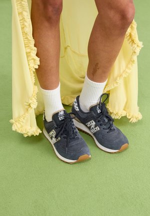 Osoba nosząca czarne buty sportowe New Balance z detalami w panterkę, białe skarpetki i żółtą falbaniaste sukienkę na zielonej podłodze.
