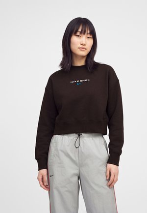 Ung kvinde med glat sort hår bærer en sort Nike Shox sweatshirt og lysegrå snørrebukser med røde striber.