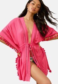 Copertura rosa con maniche corte, dotata di una cintura legata in vita, dettagli in crochet e pompon lungo i bordi. Tessuto leggero.