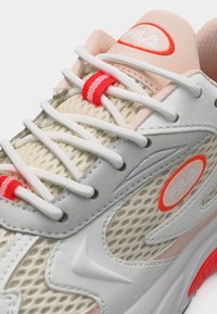 Κοντινό πλάνο μπεζ και άσπρου αθλητικού παπουτσιού FILA με ύφασμα από δίχτυ, άσπρα κορδόνια, κόκκινα σημεία και λογότυπο στη γλώσσα και στο πλάι.