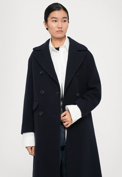 Donna che indossa un lungo cappotto doppiopetto blu scuro sopra una camicia bianca e jeans blu, che guarda a sinistra su uno sfondo semplice.