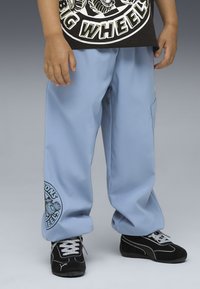 Niño lleva pantalones cargo azul claro con un gráfico en una pierna, una camiseta negra de "Big Wheels" y zapatillas Puma negras con cordones blancos.