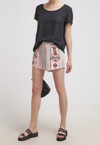 Donna con una maglietta nera a maniche corte, pantaloni corti a fantasia con design geometrici, sandali neri e in mano una borsa nera, su uno sfondo semplice.