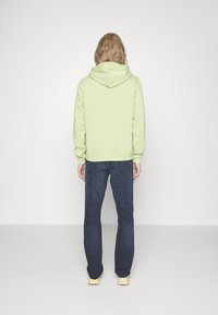 Sudadera verde claro con un bolsillo trasero, capucha ajustable y puños acanalados, combinada con jeans azul oscuro y zapatillas con tintes amarillos.