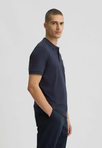 Polo de color navy con mangas cortas y cuello, que cuenta con un bolsillo y sutiles acentos de puntos a lo largo del cuello. Textura de tela suave.