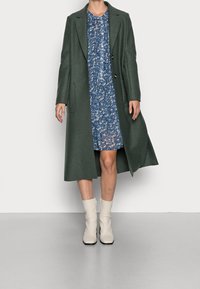 Langer grüner Wollmantel mit Reverskragen und zwei Knöpfen, kombiniert mit einem blauen Blumenkleid und weißen Stiefeletten. Glatte Textur.