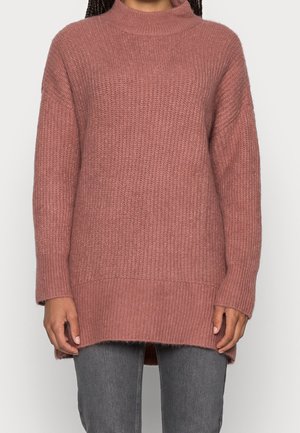 Personne portant un long pull tricoté mauve avec un col haut et un ourlet côtelé, associé à un pantalon gris sur fond blanc.