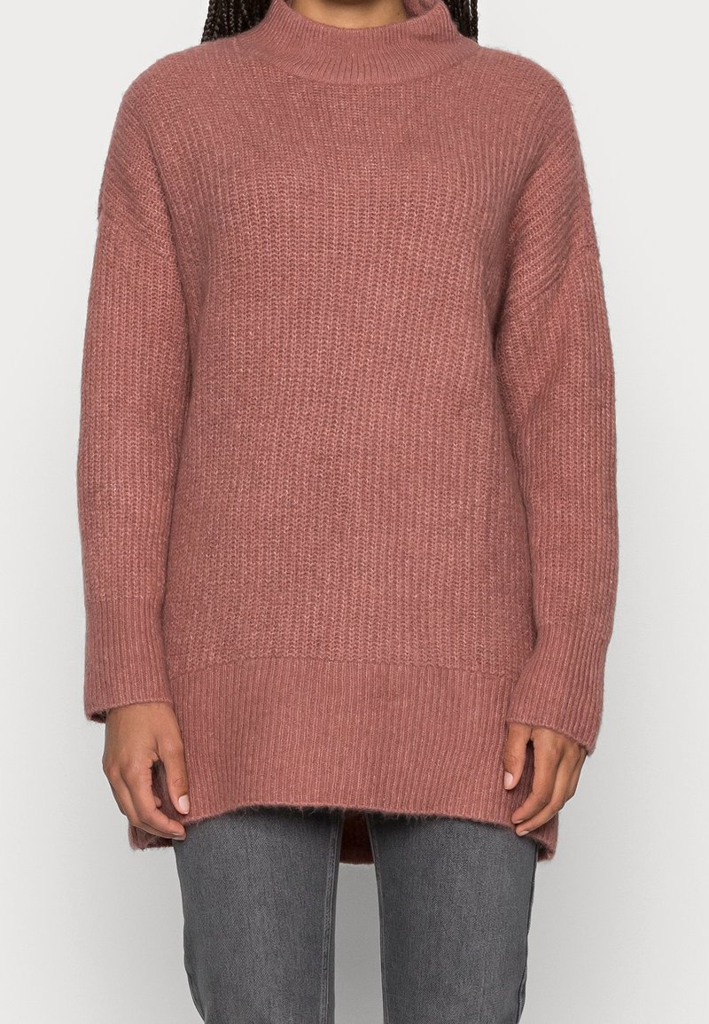 Personne portant un long pull tricoté mauve avec un col haut et un ourlet côtelé, associé à un pantalon gris sur fond blanc.