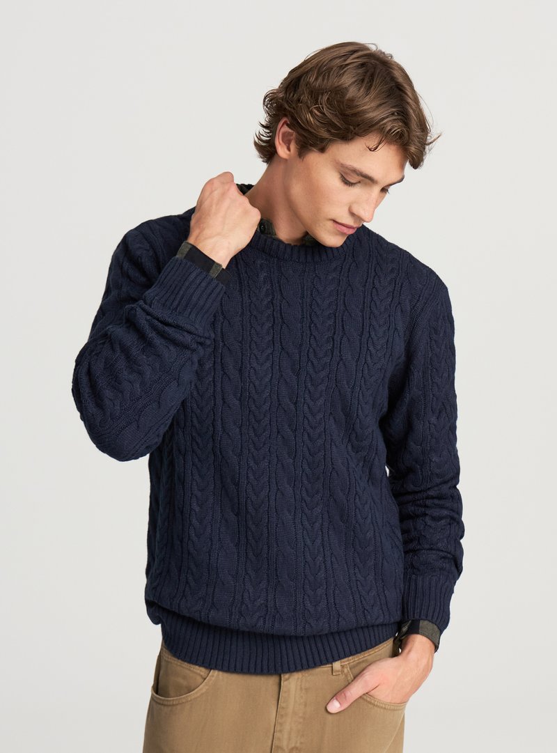 Maglione blu navy a trecce con scollo rotondo e polsini a coste. Indossato sopra una camicia a quadri con colletto, abbinato a pantaloni beige.