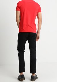 Czerwona koszulka z krótkim rękawem w zestawie z czarnymi spodniami z podwiniętymi mankietami i czarnymi butami. Materiał wydaje się gładki i dopasowany.