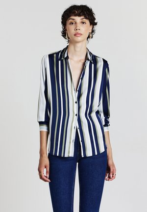Jeune femme avec des cheveux courts et bouclés portant des boucles d'oreilles en or, une chemise à rayures verticales bleues, vertes et blanches, et un jean bleu foncé.
