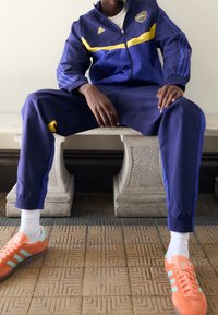 Modrý sportovní komplet se žlutými detaily, zipová bunda, elastické manžety a odpovídající kalhoty. Růžovo-oranžové boty se zelenými pruhy. Bílé ponožky.