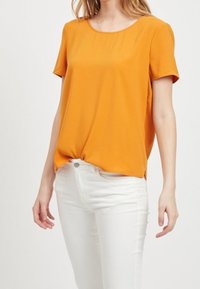 Short-sleeve, round-neck top in vibrant orange fabric, featuring a relaxed fit and side slits, paired with white jeans.