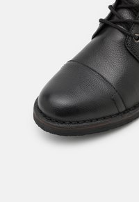 Black leather shoe with a textured surface, round toe, stitched detailing along the sole, and a contrasting brown accent.