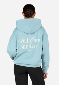 Ανοιχτό μπλε φούτερ με ευρύχωρη κουκούλα. Στην πλάτη αναγράφεται το κείμενο "Self Care Sundays." σε κρέμα χρώμα. Μαλακό ύφασμα, χαλαρή εφαρμογή.