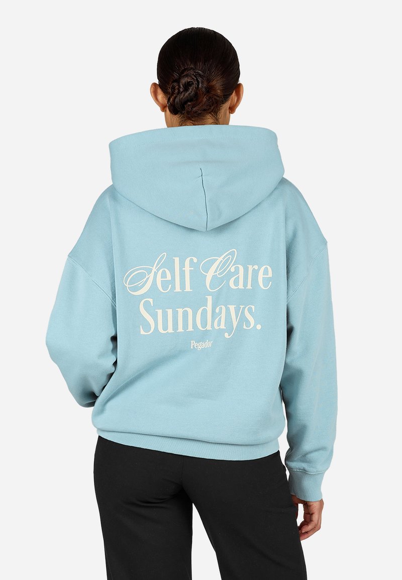 Ανοιχτό μπλε φούτερ με ευρύχωρη κουκούλα. Στην πλάτη αναγράφεται το κείμενο "Self Care Sundays." σε κρέμα χρώμα. Μαλακό ύφασμα, χαλαρή εφαρμογή.