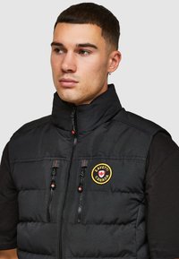 Zwarte gewatteerde vest met hoge kraag, twee ritsborstzakken, rode stikaccenten en een cirkelvormig geborduurd logo aan de linkerkant.