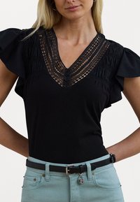 Μαύρη τοπ με V λαιμόκοψη και δαντελένιες λεπτομέρειες, με κοντά φρου-φρου μανίκια, συνδυασμένη με ανοιχτό μπλε τζιν ψηλόμεσο και μαύρη ζώνη με ασημένιες λεπτομέρειες.