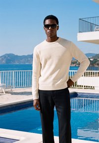 Homme portant des lunettes de soleil noires, un pull crème texturé et un pantalon noir, debout près d'une piscine avec l'océan et des montagnes en arrière-plan.