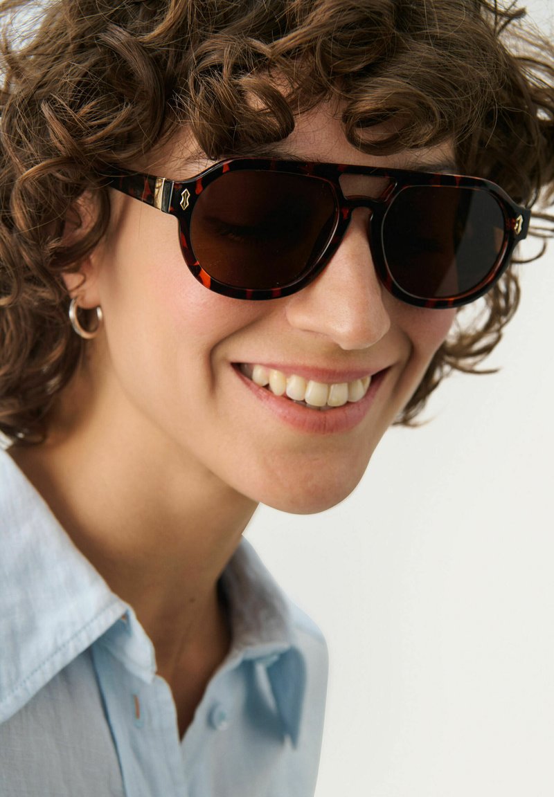 Tortoiseshell sunglasses with dark lenses, rectangular shape, and gold accents. Model wears a light blue collared shirt, smiling.