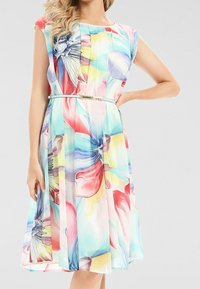 Kvinna som bär en ärmlös flerfärgad blommig klänning med veck och ett tunt ljusblått bälte, stående med ena handen på höften.