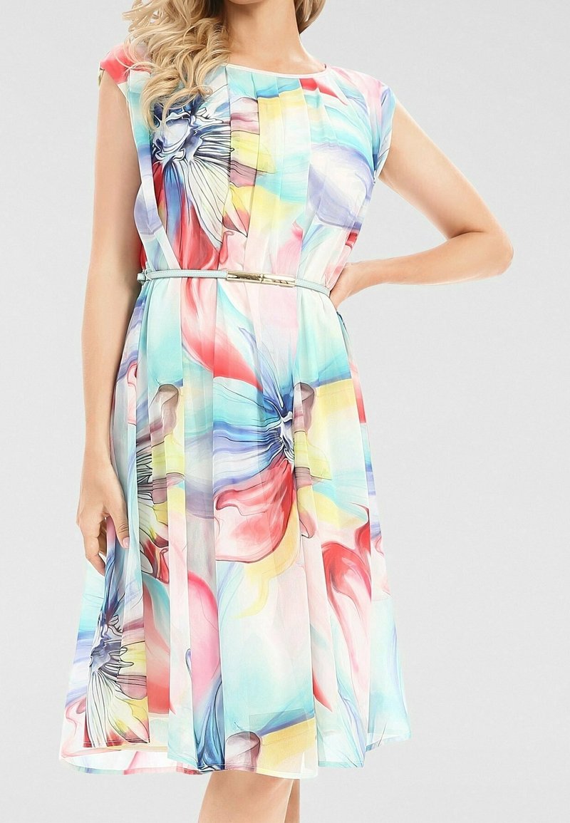 Kvinna som bär en ärmlös flerfärgad blommig klänning med veck och ett tunt ljusblått bälte, stående med ena handen på höften.