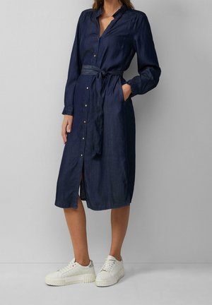 Kobieta w ciemnoniebieskiej midi sukience z guzikami, długimi rękawami i paskiem w talii, w białych sneakersach na platformie, stojąca na tle szarej ściany.