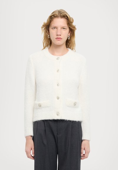 Weißer, kurzer Cardigan aus flauschigem Strick mit langen Ärmeln, rundem Halsausschnitt und dekorativen Knöpfen. Verfügt über zwei Fronttaschen.