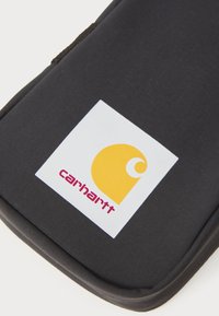 Schwarzes Stofftäschchen mit einem auffälligen weißen Etikett, das ein gelbes Logo und roten Text mit der Aufschrift "carhartt" zeigt, sanft genähte und konturierte Kanten.