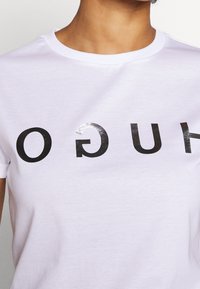 Camiseta de algodón blanca con cuello redondo, que presenta texto negro brillante que dice "HUGO" en una fuente sans-serif en negrita. Mangas cortas visibles.