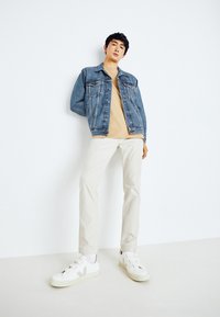 Chaqueta de mezclilla con dos bolsillos en el pecho, camiseta beige, pantalones ajustados crema y zapatillas blancas sin cordones. Fondo plano.